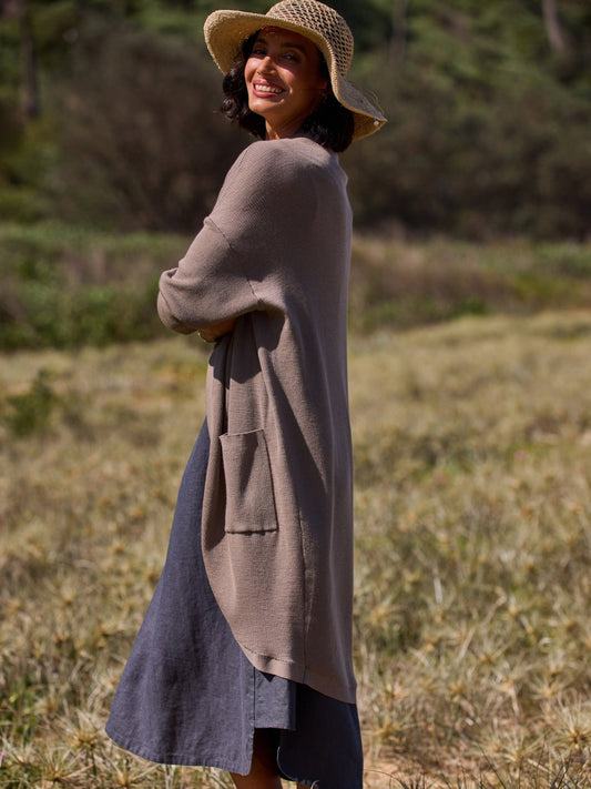 Woman wearing a long brown coat and straw hat standing in a natural setting.