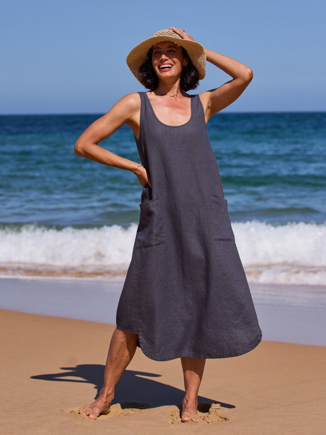Woman in a drak dress and straw hat standing on a beach with ocean waves in the background