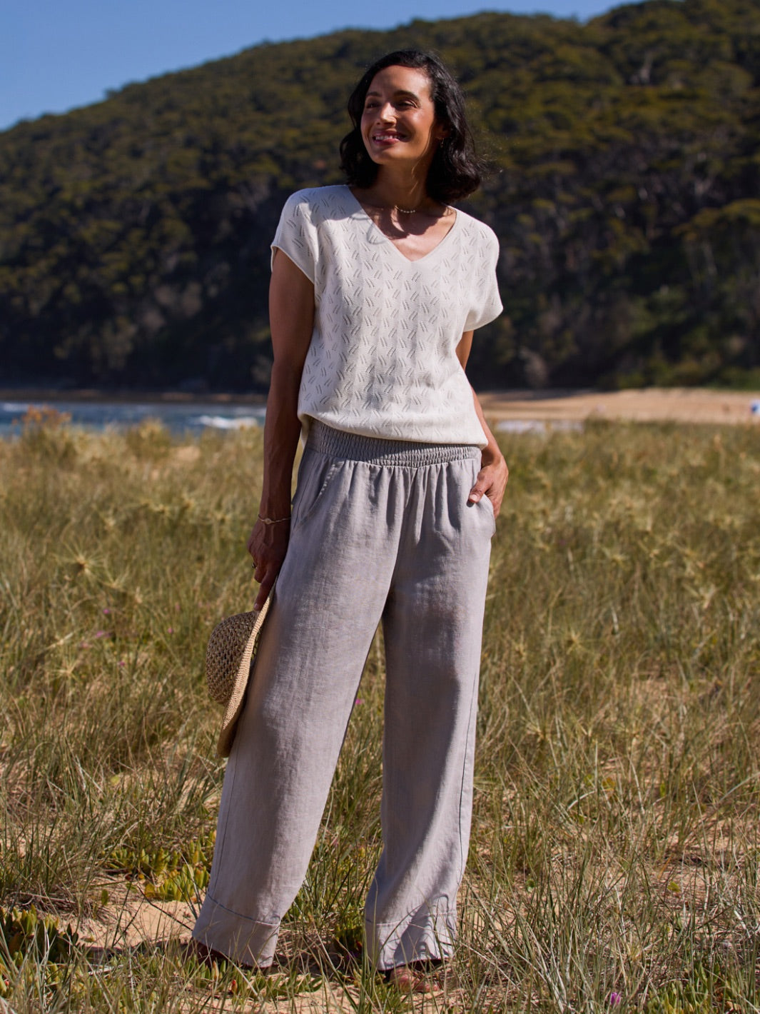 Woman standing in a natural setting wearing a light top and gray pants.