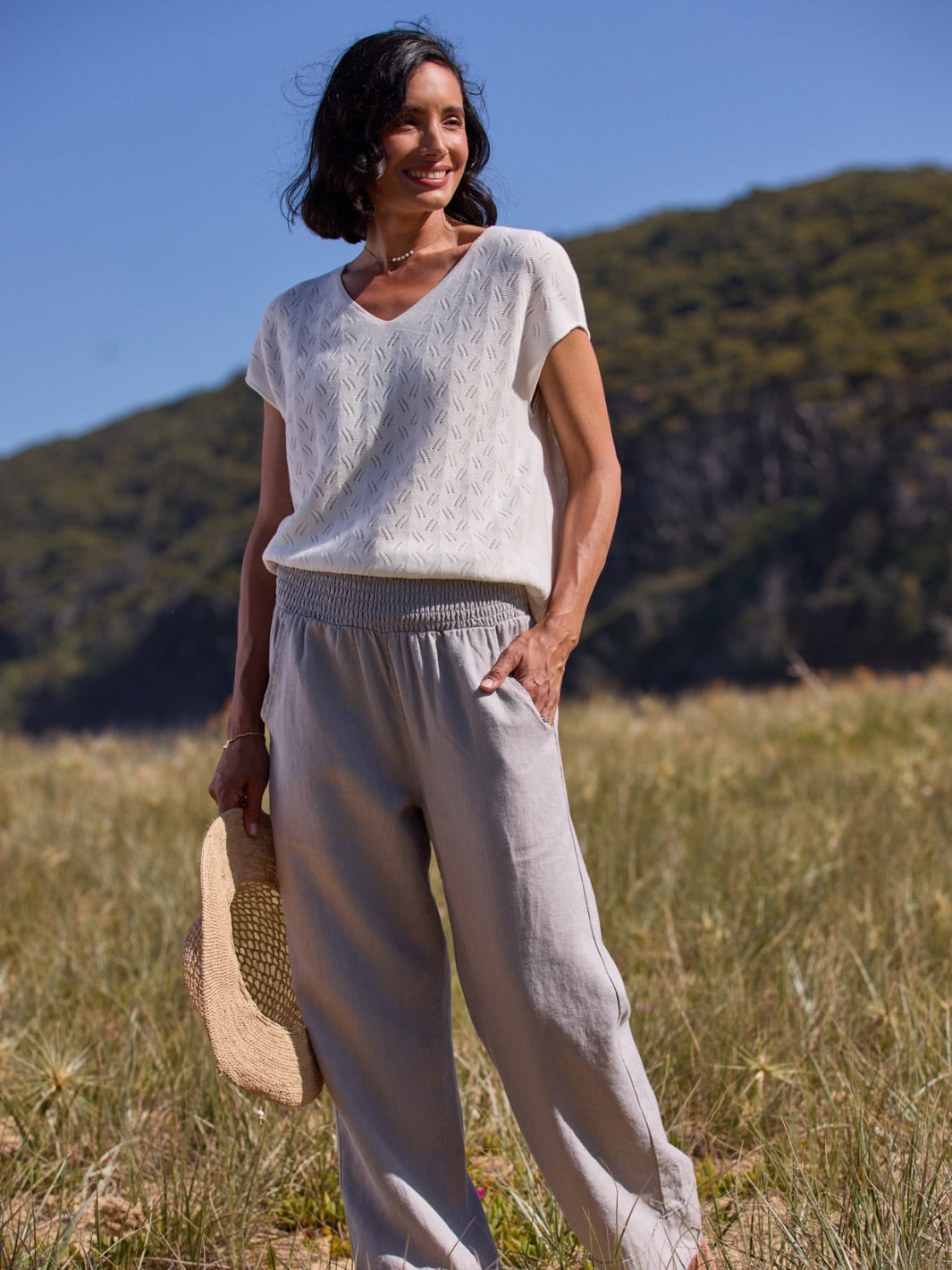 Woman in a white top and light gray pants standing in a field with mountains in the background