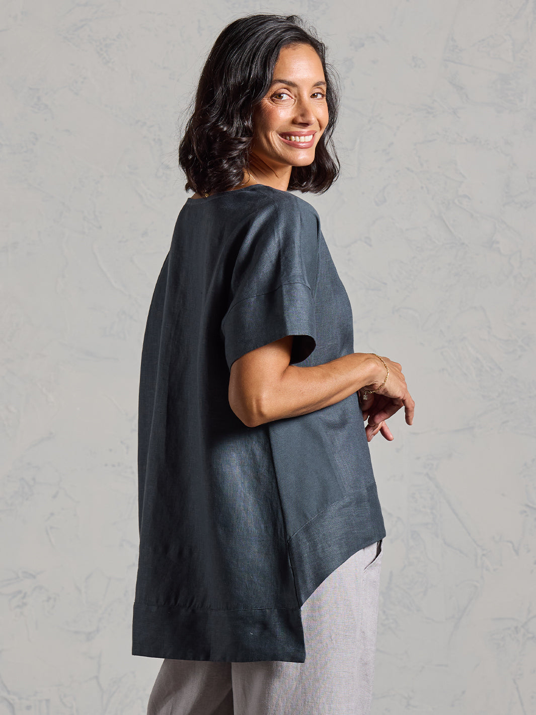 Woman wearing a dark gray tunic over light pants against a plain background