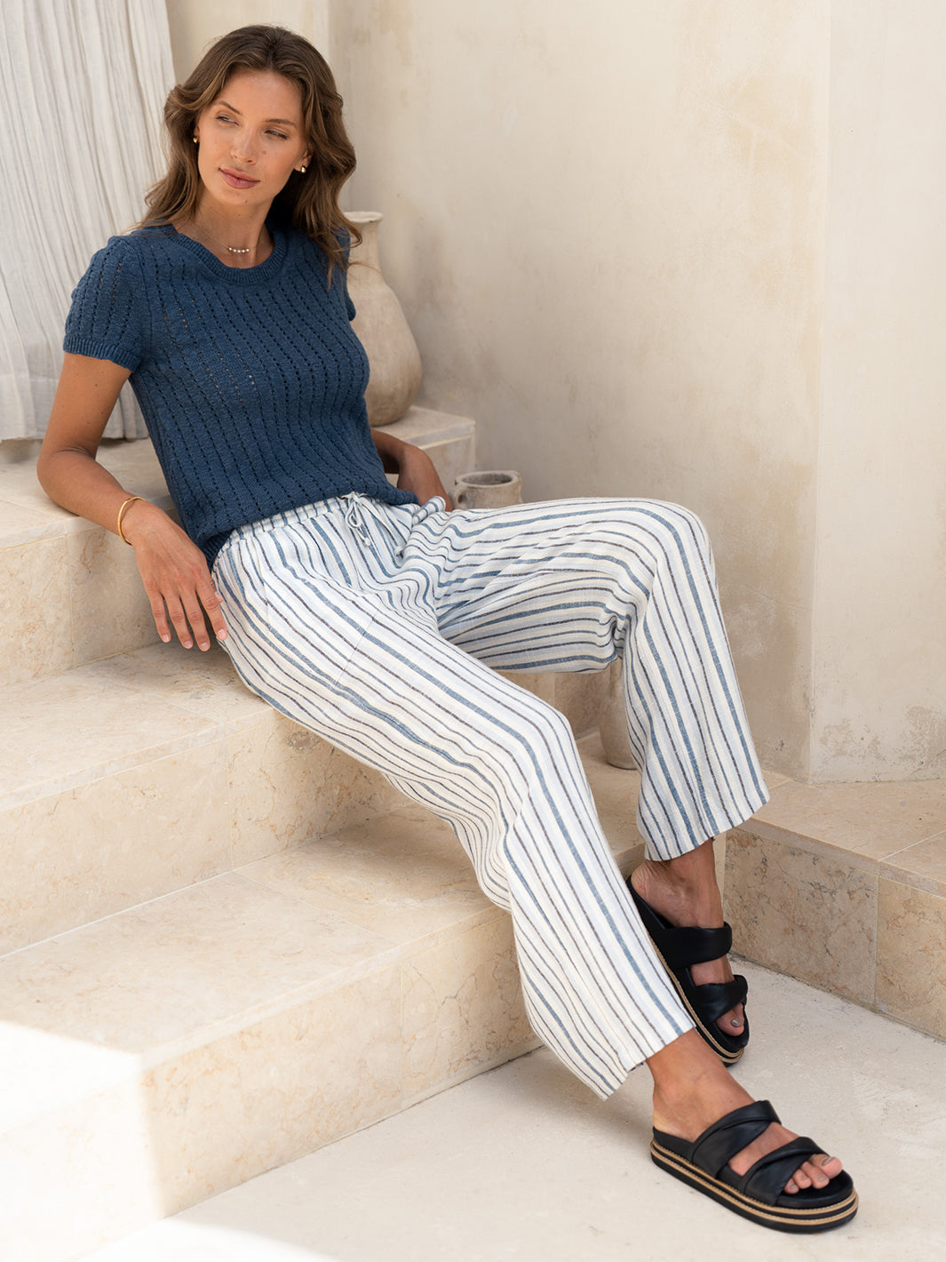 Model seated in the Jolie Linen Blend Trousers in coastal blue with a navy knit top, on sunlit stone steps.