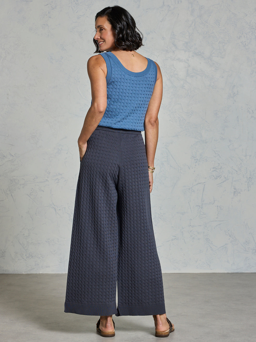 Woman wearing a blue sleeveless top and dark gray wide-leg pants against a plain background
