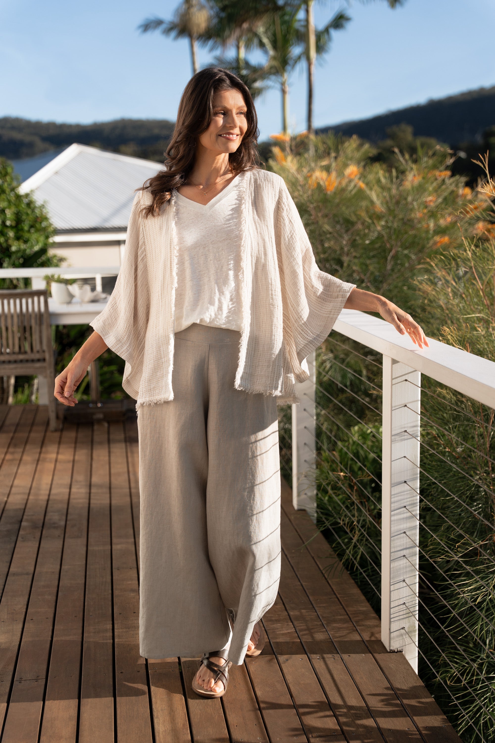 Woman in a white outfit standing on a wooden deck with greenery and a house in the background