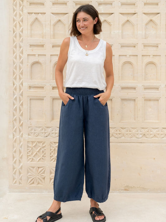 Woman wearing a white sleeveless top and blue wide-leg pants standing against a decorative stone wall.