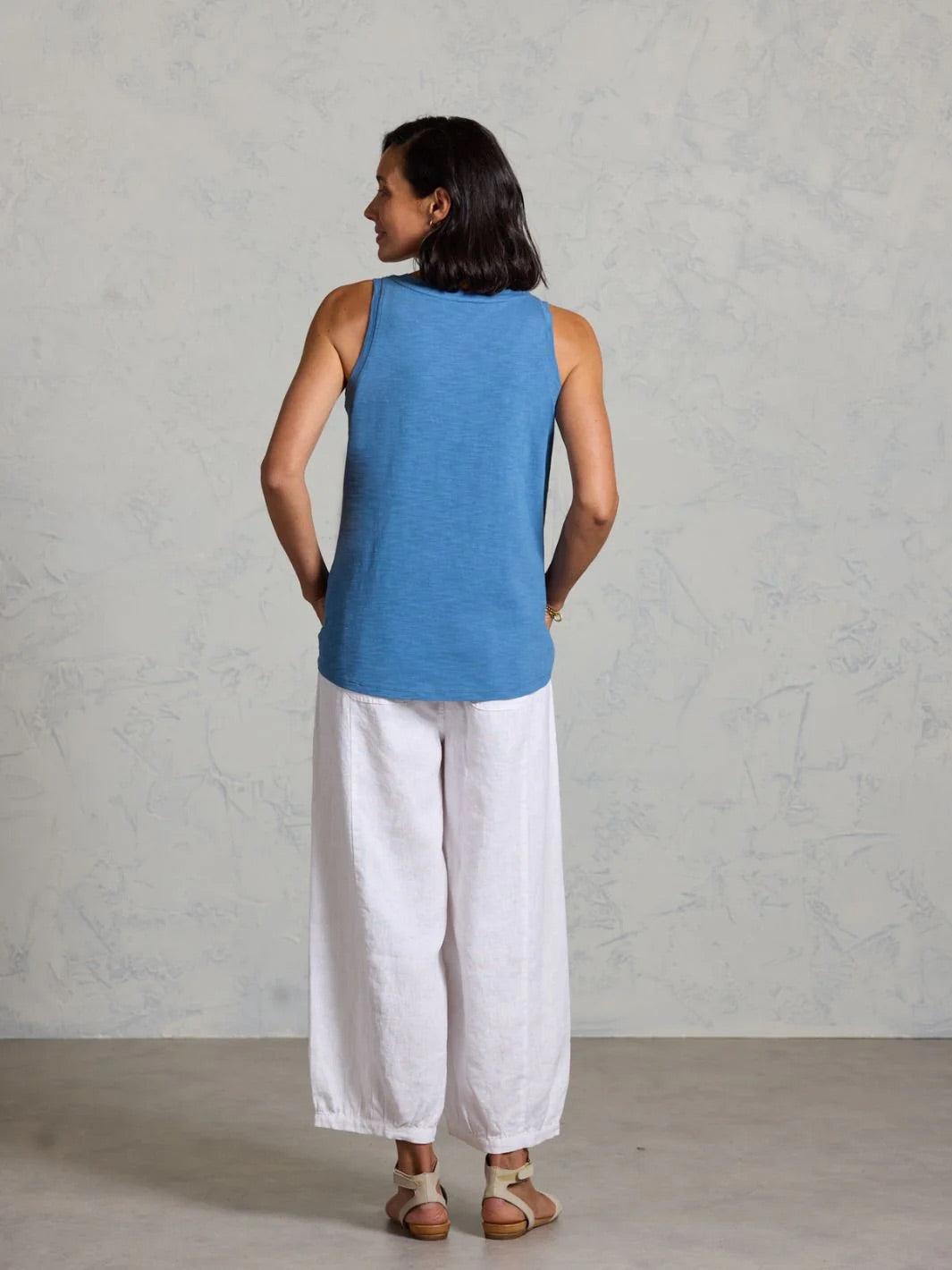 Woman wearing a blue tank top and white pants on a grey background.