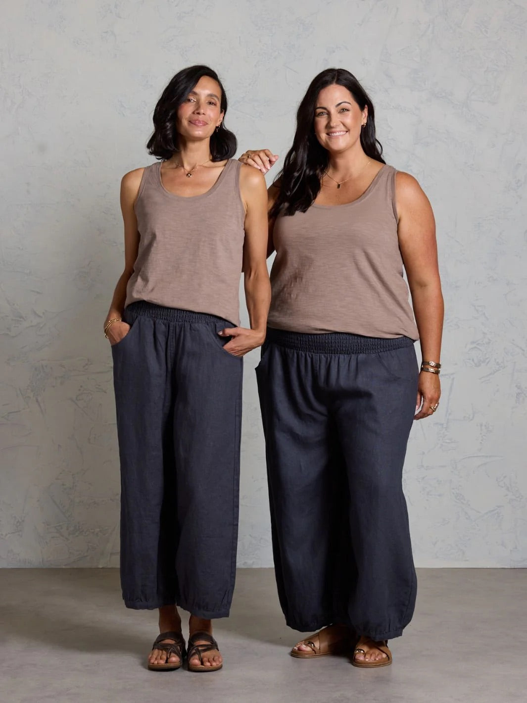 Two women wearing matching brown tank tops and black pants standing against a plain background.