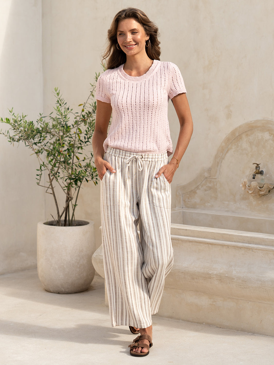 Model wearing the Jolie Linen Blend Trousers in driftwood with a pink knit top, beside an olive tree