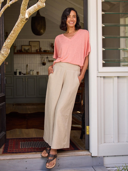 Woman standing at the door of a house wearing a pink top and beige pants.