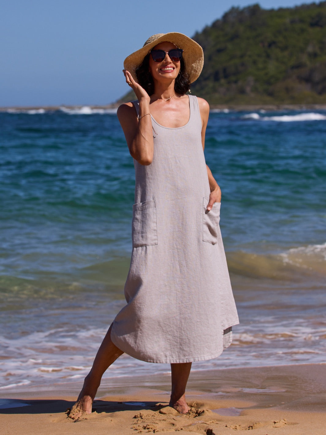 Woman in a gray dress and sun hat standing on a beach with ocean waves in the background