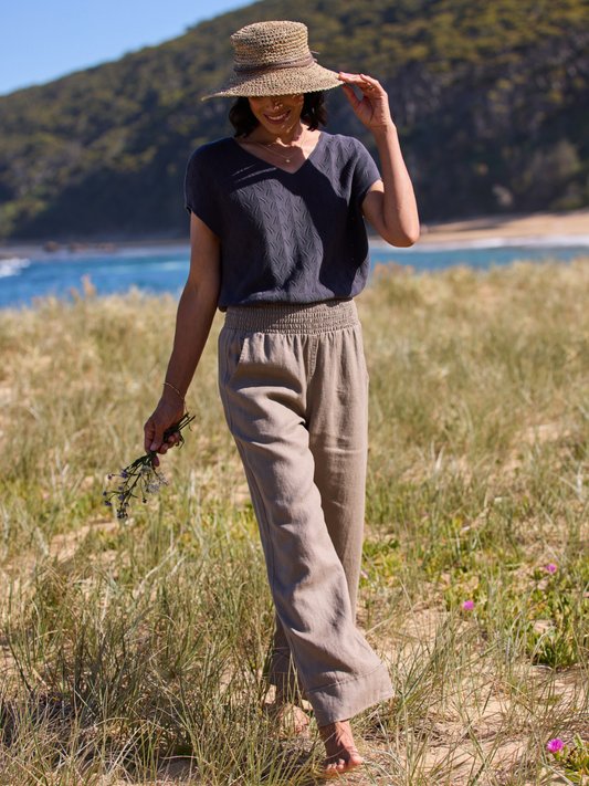 Woman in a natural setting wearing a straw hat, dark top, and beige pants.