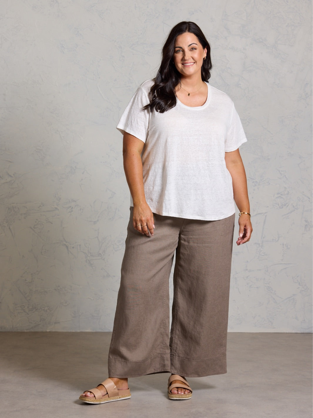 Woman wearing a white top and brown pants against a gray background