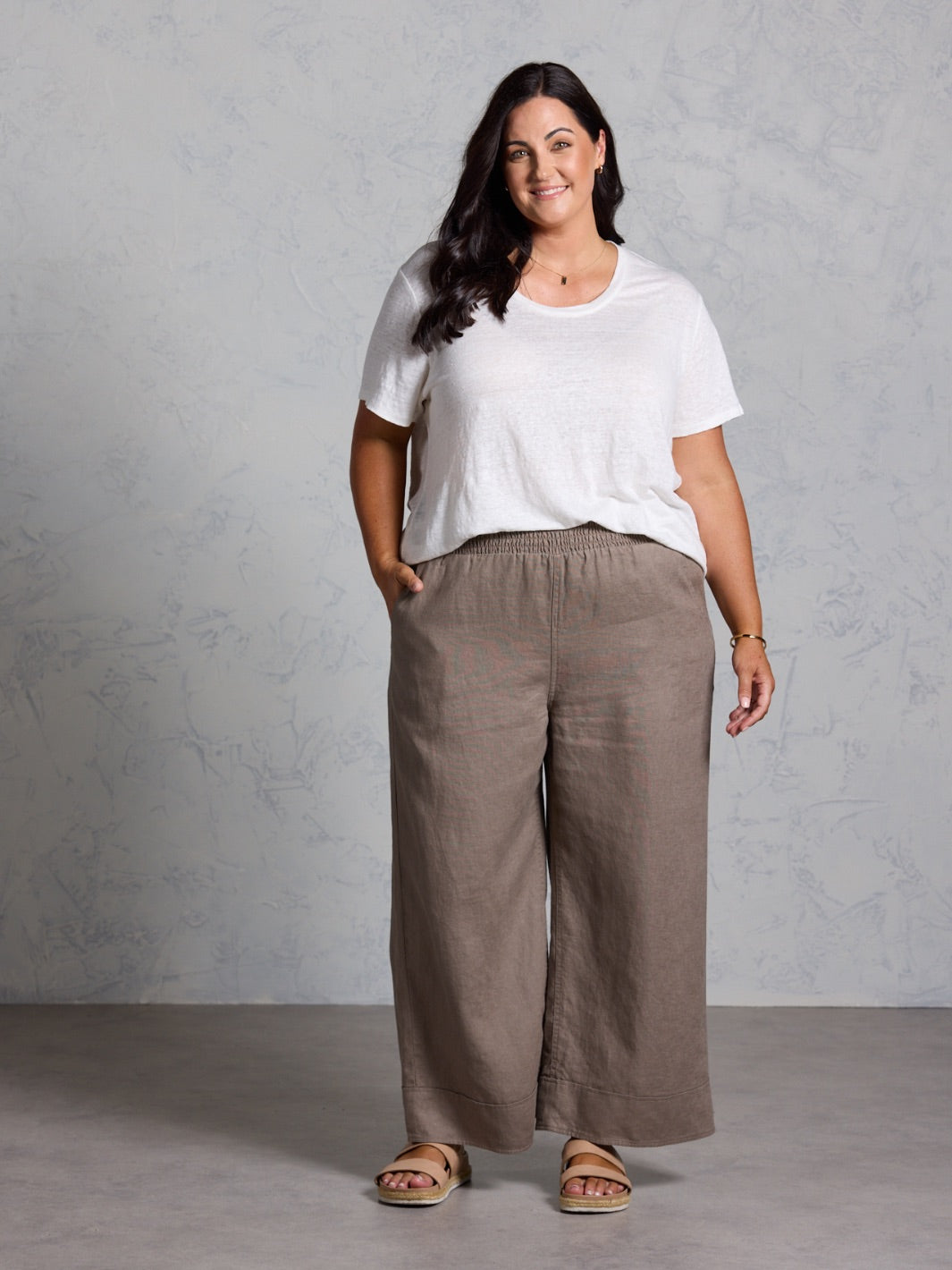 Woman wearing a white t-shirt and brown pants against a gray background