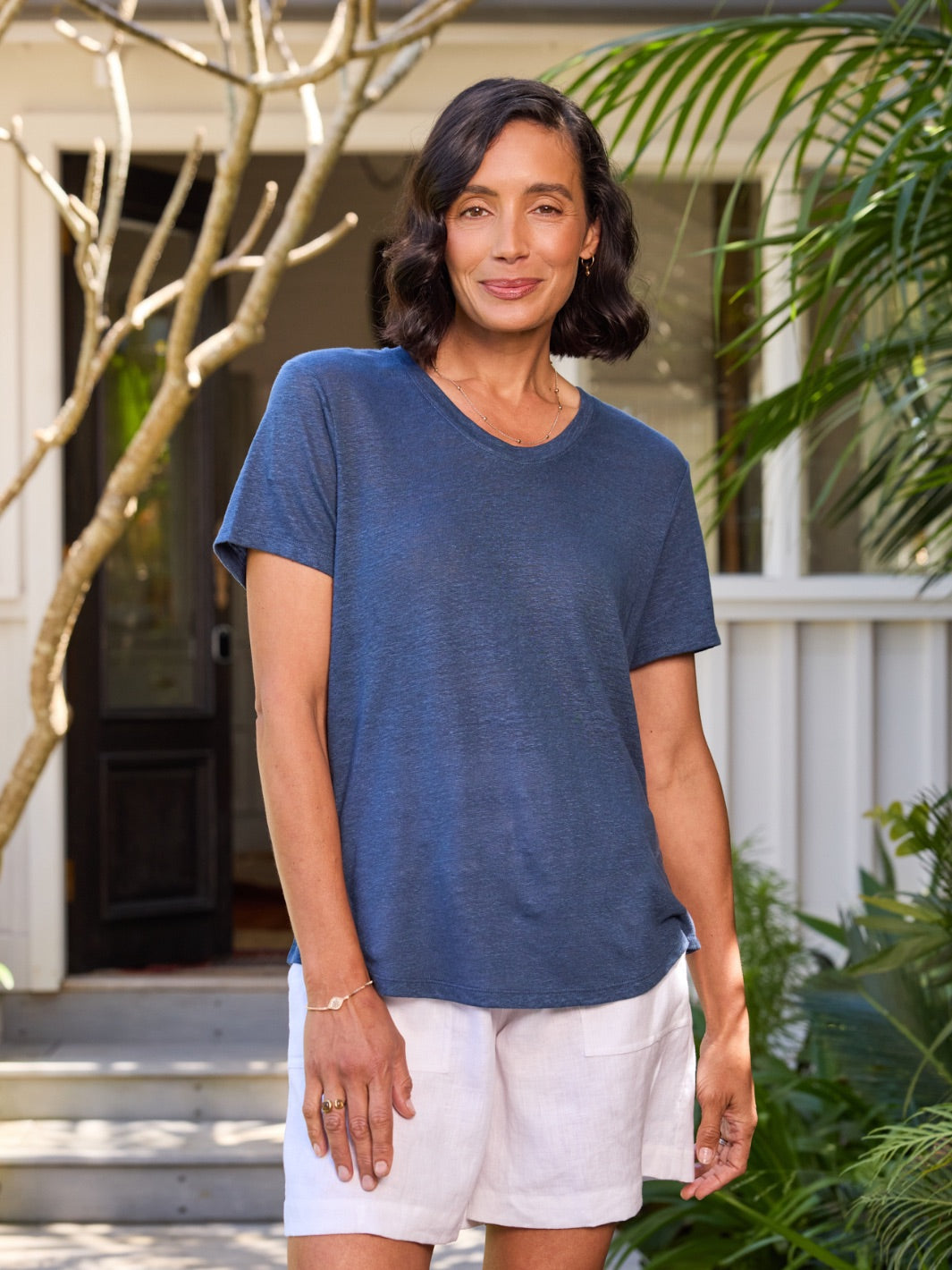 Woman wearing a blue top and white shorts standing outdoors with plants and a building in the background