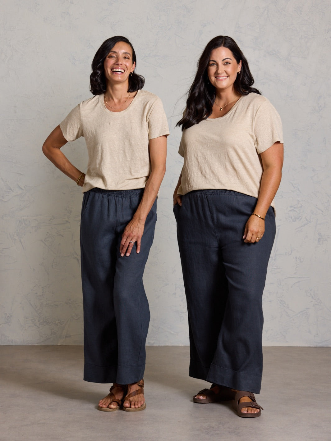 Two women wearing matching outfits of beige tops and black pants against a plain background.