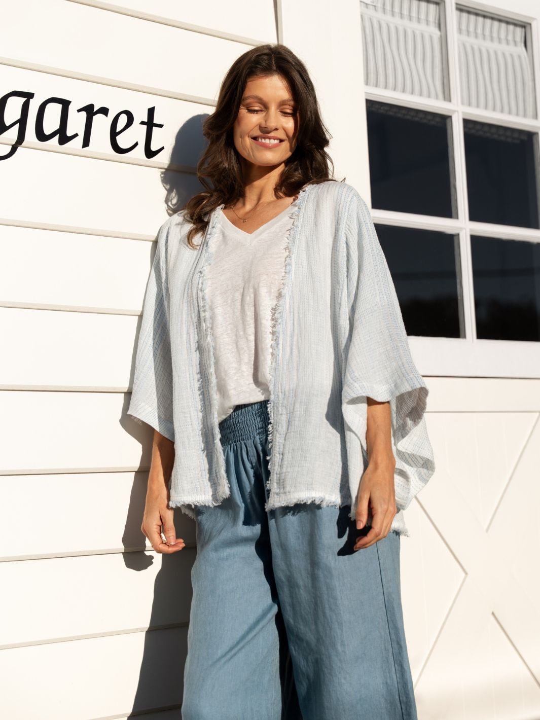 Woman wearing a light blue kimono-style garment and blue pants standing in front of a white building.
