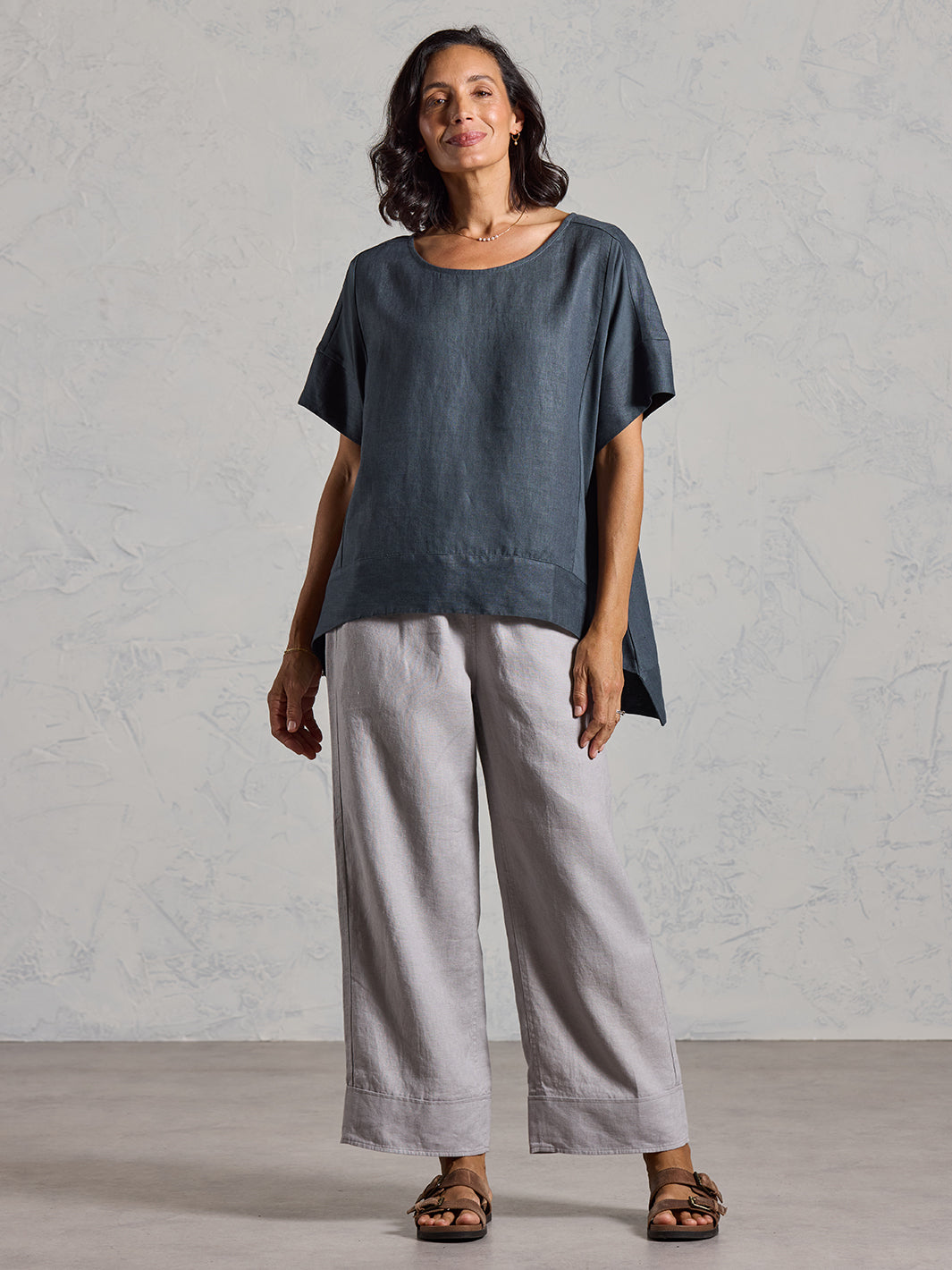 Woman wearing a dark top and light gray pants against a plain background