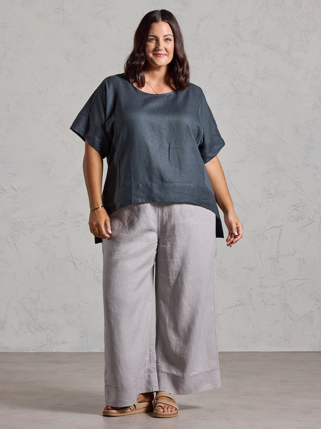 Woman wearing a dark top and light gray pants against a plain background