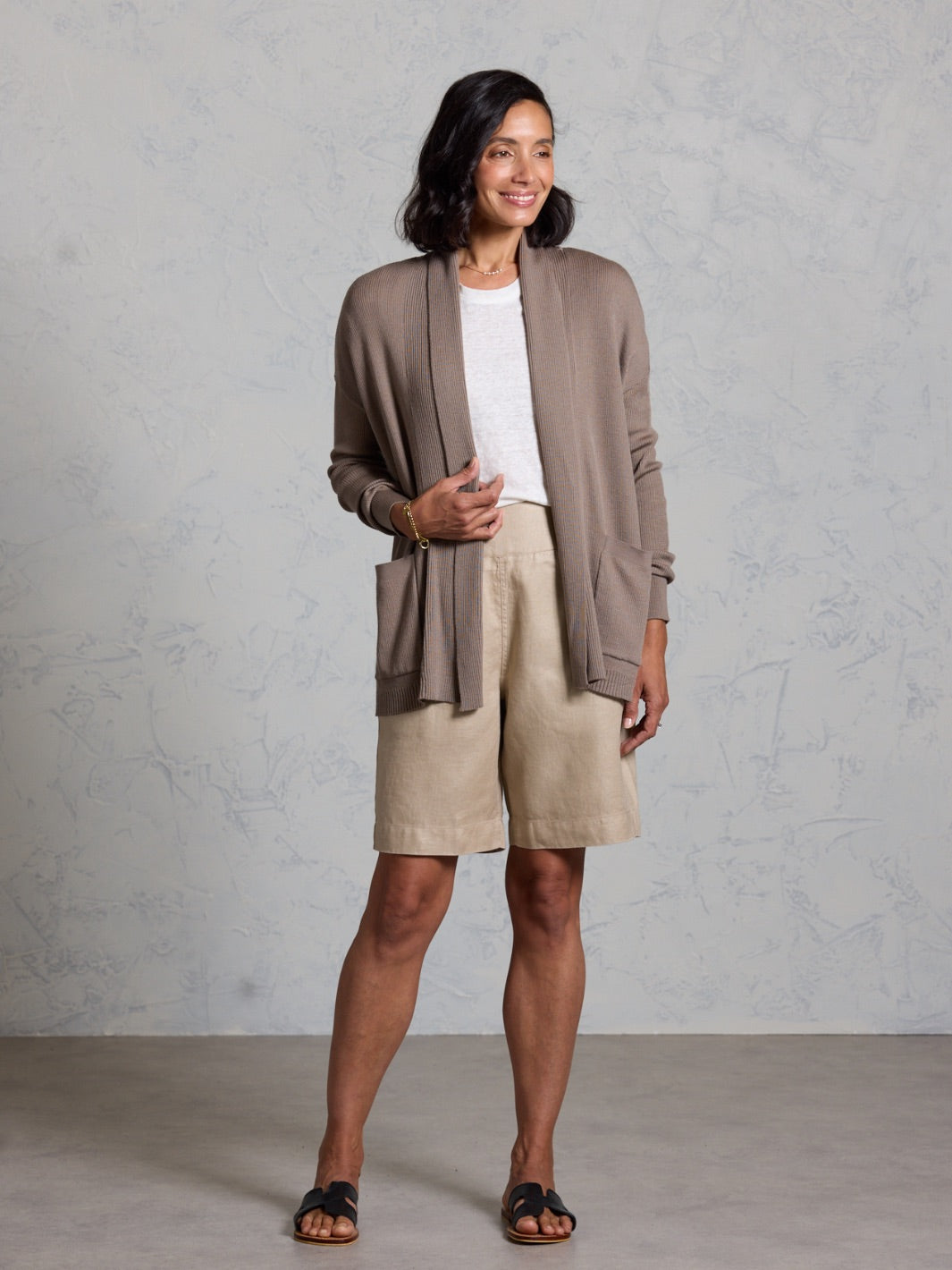 Woman wearing a brown cardigan and beige shorts against a gray background