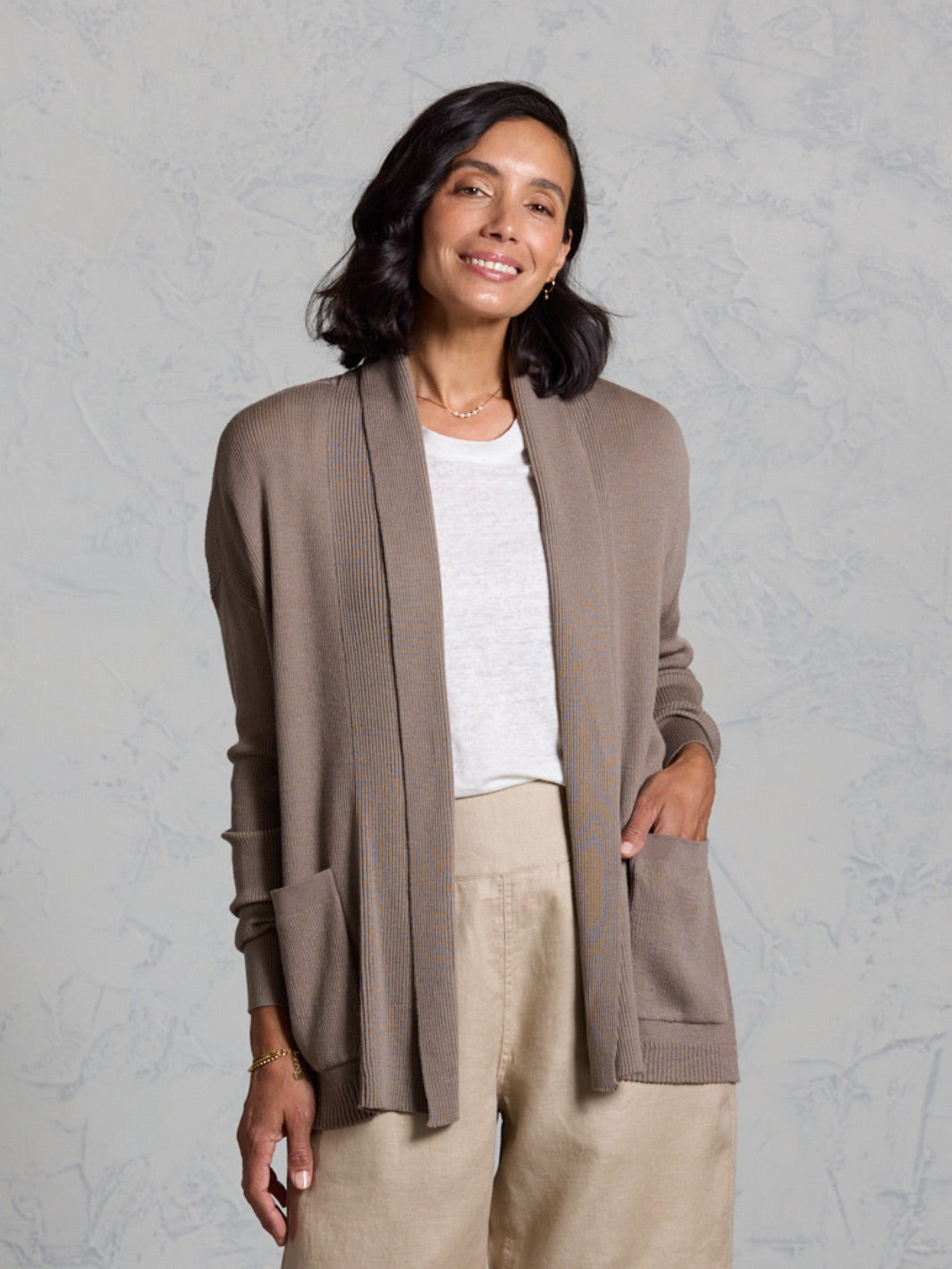 Woman wearing a brown cardigan over a white shirt and beige pants against a gray background