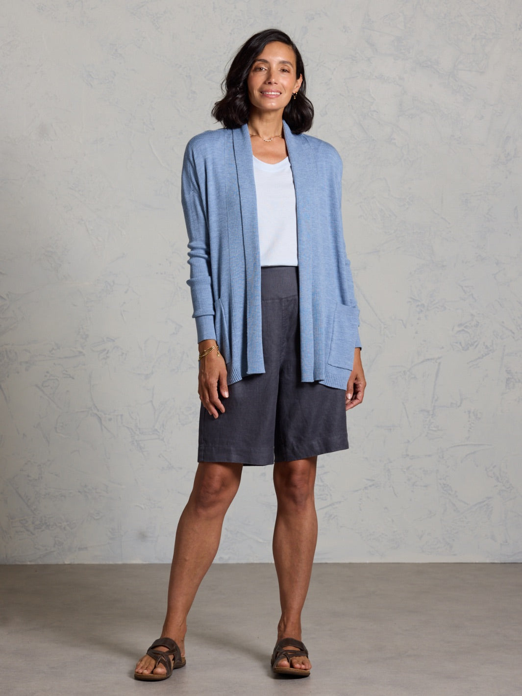 Woman wearing a blue cardigan over a white shirt and dark shorts against a gray background