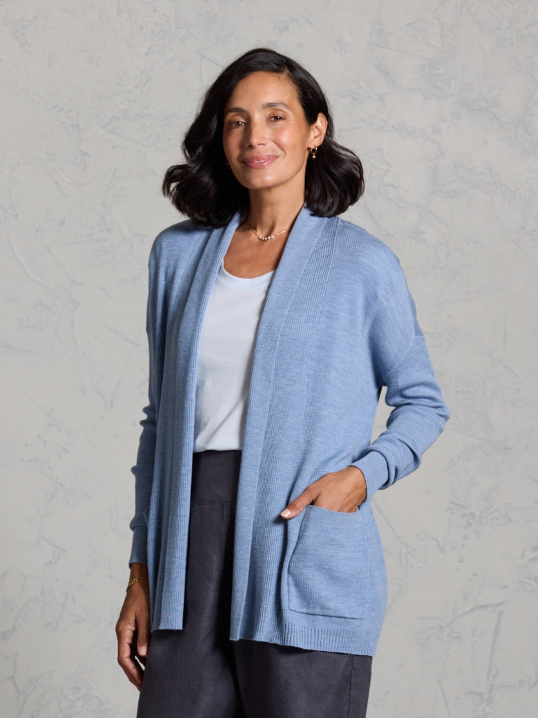 Woman wearing a light blue cardigan over a white shirt and dark pants on a gray background