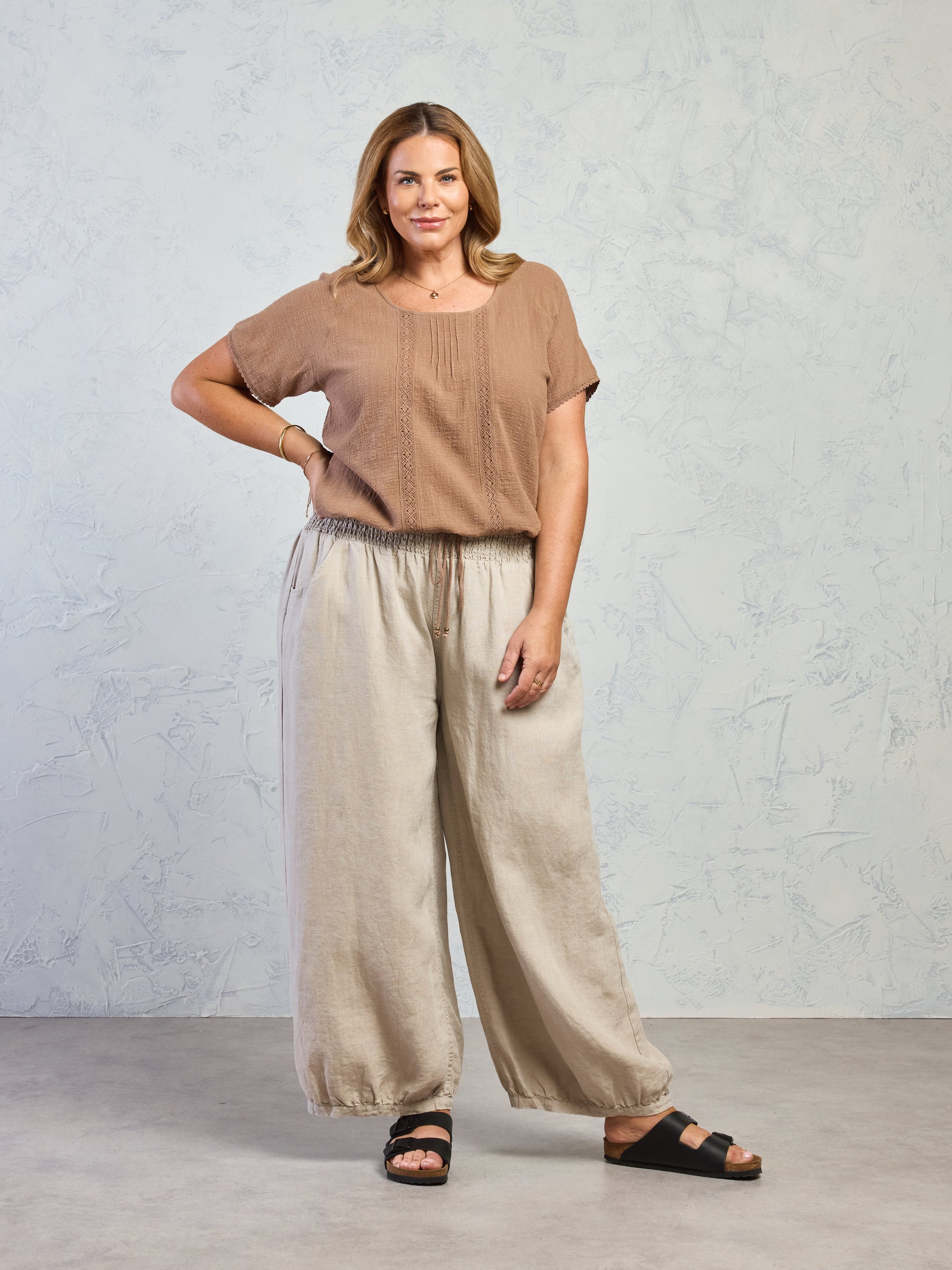 Woman wearing a brown top and beige wide-leg pants against a light gray background