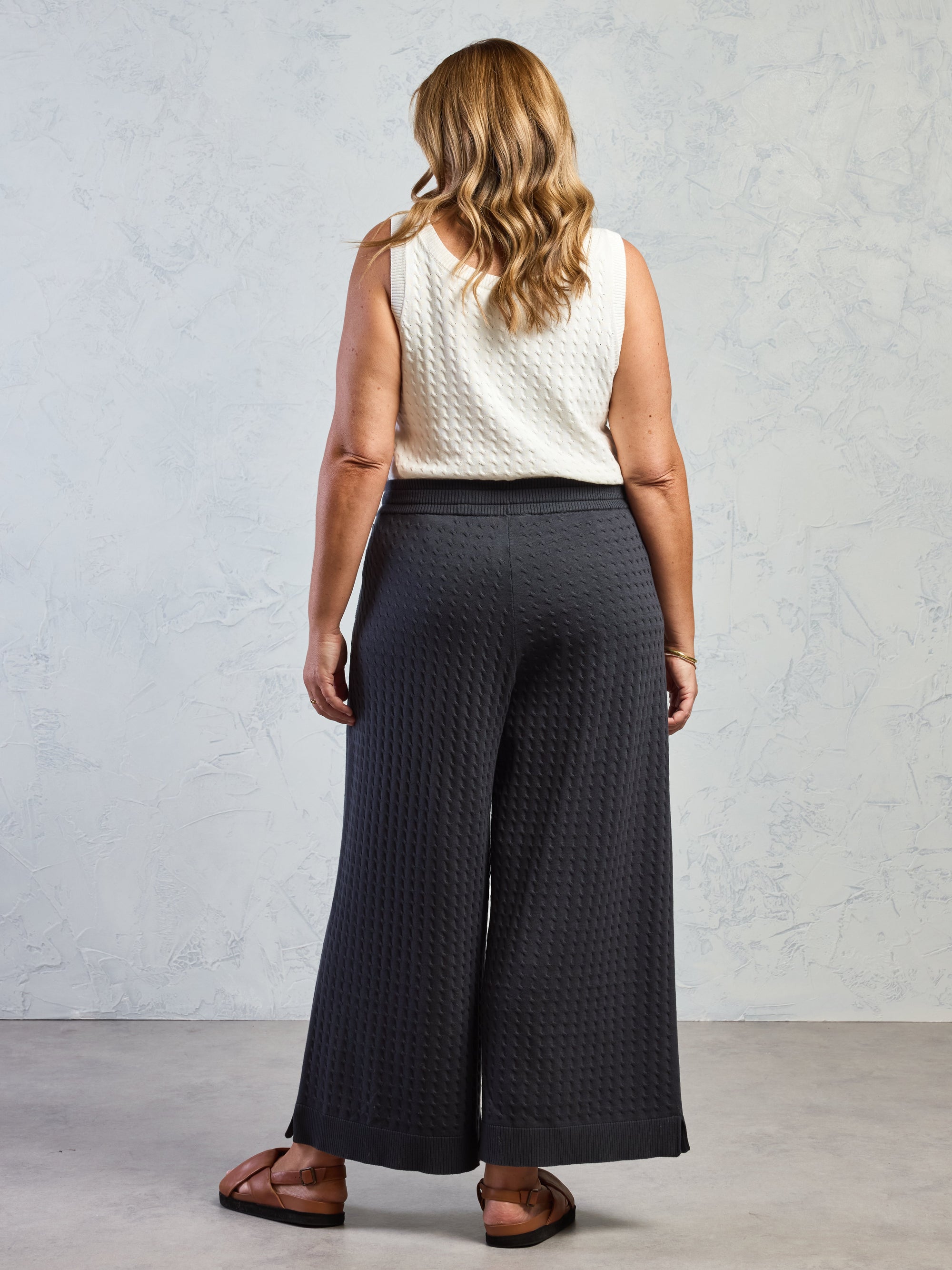 Woman wearing a white sleeveless top and dark textured pants against a gray background