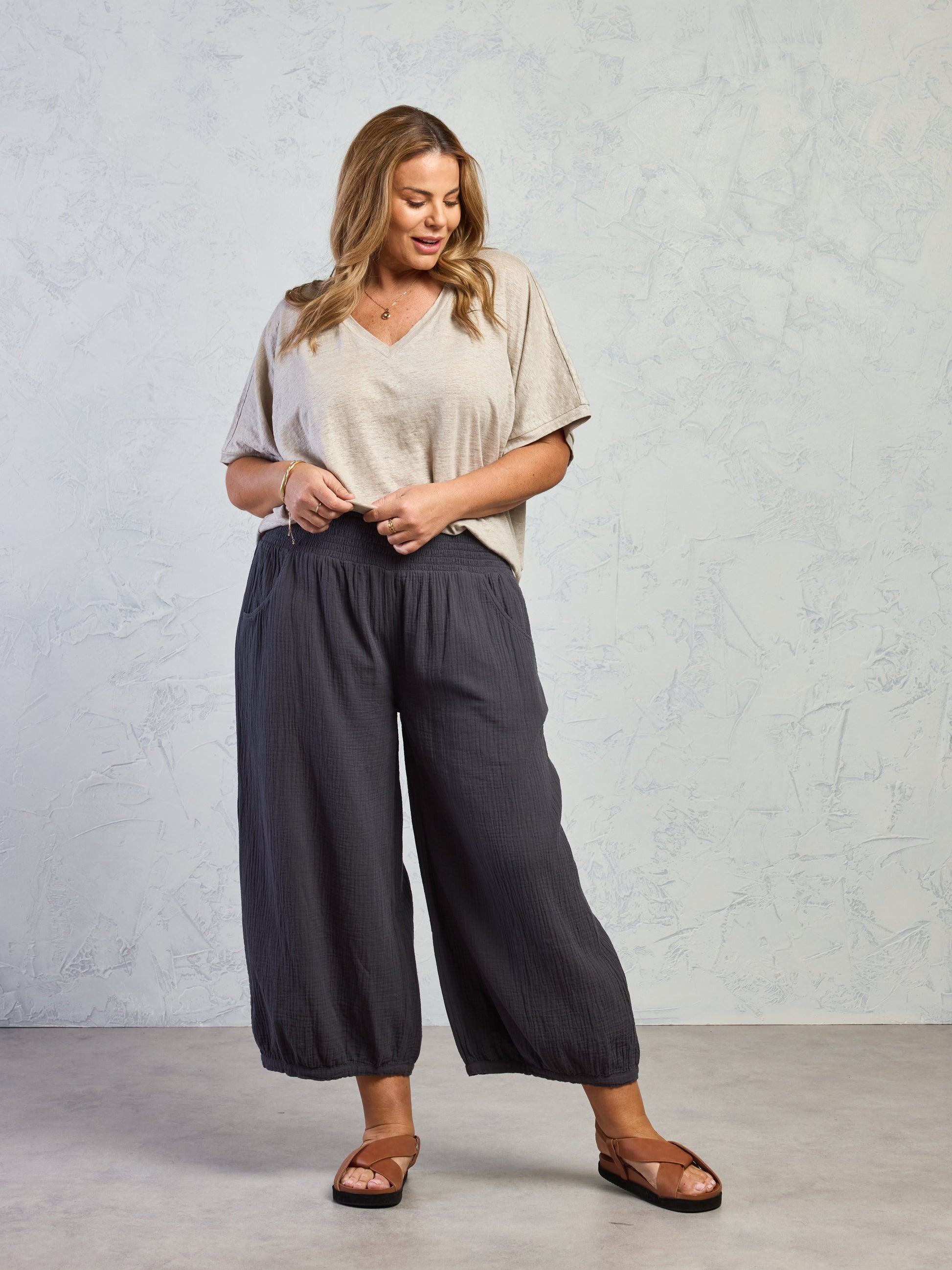 Woman wearing a beige top and dark gray wide-leg pants against a light gray background