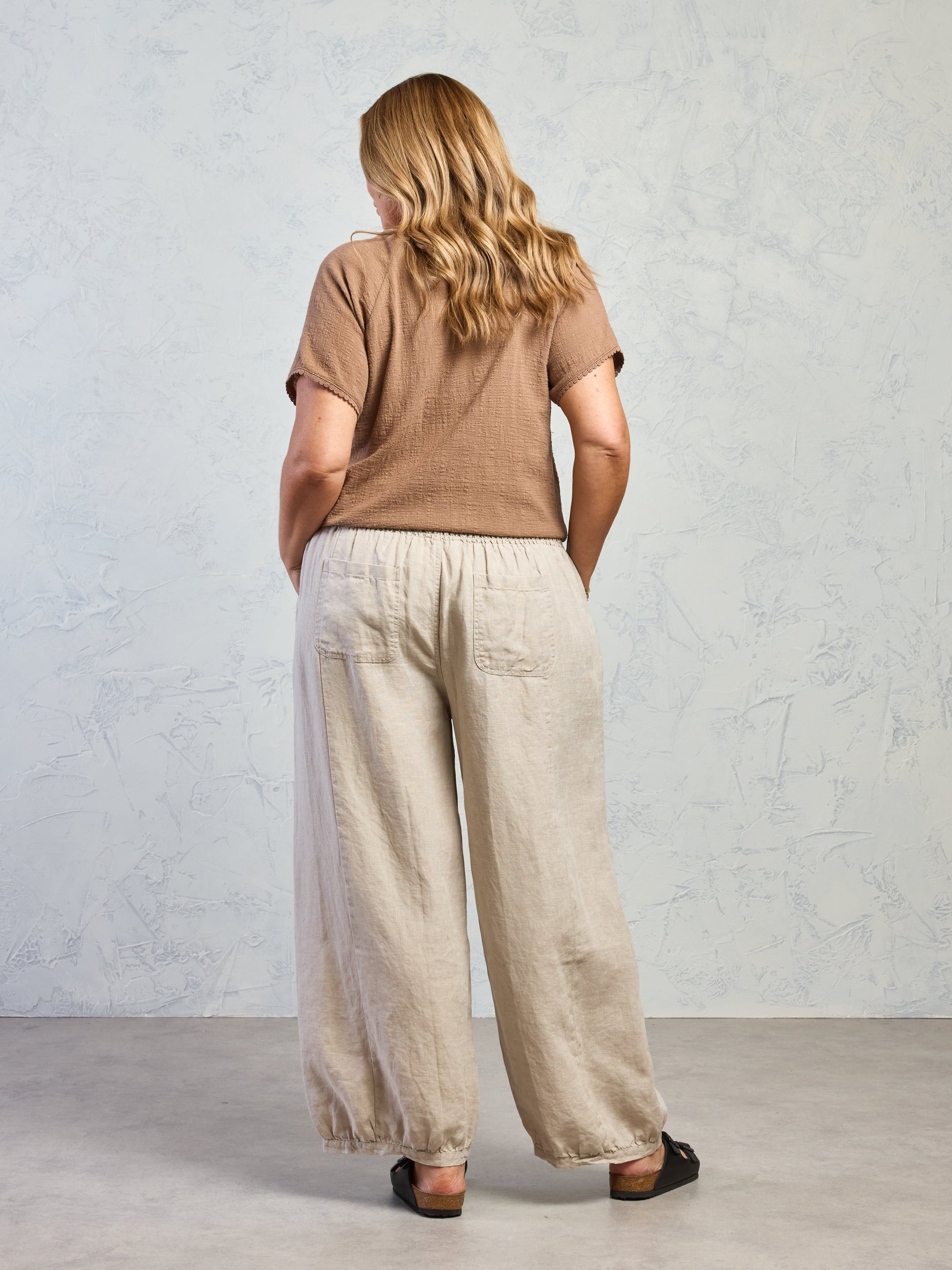 Woman wearing a brown top and beige pants against a light gray wall.