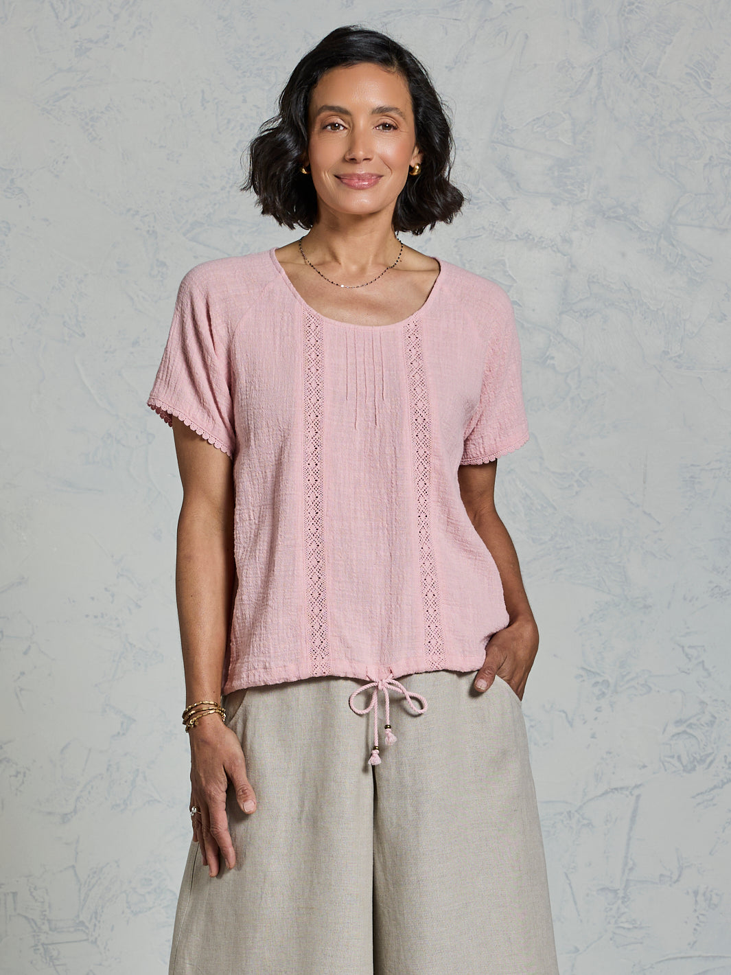 Woman wearing a pink top and beige pants against a gray background