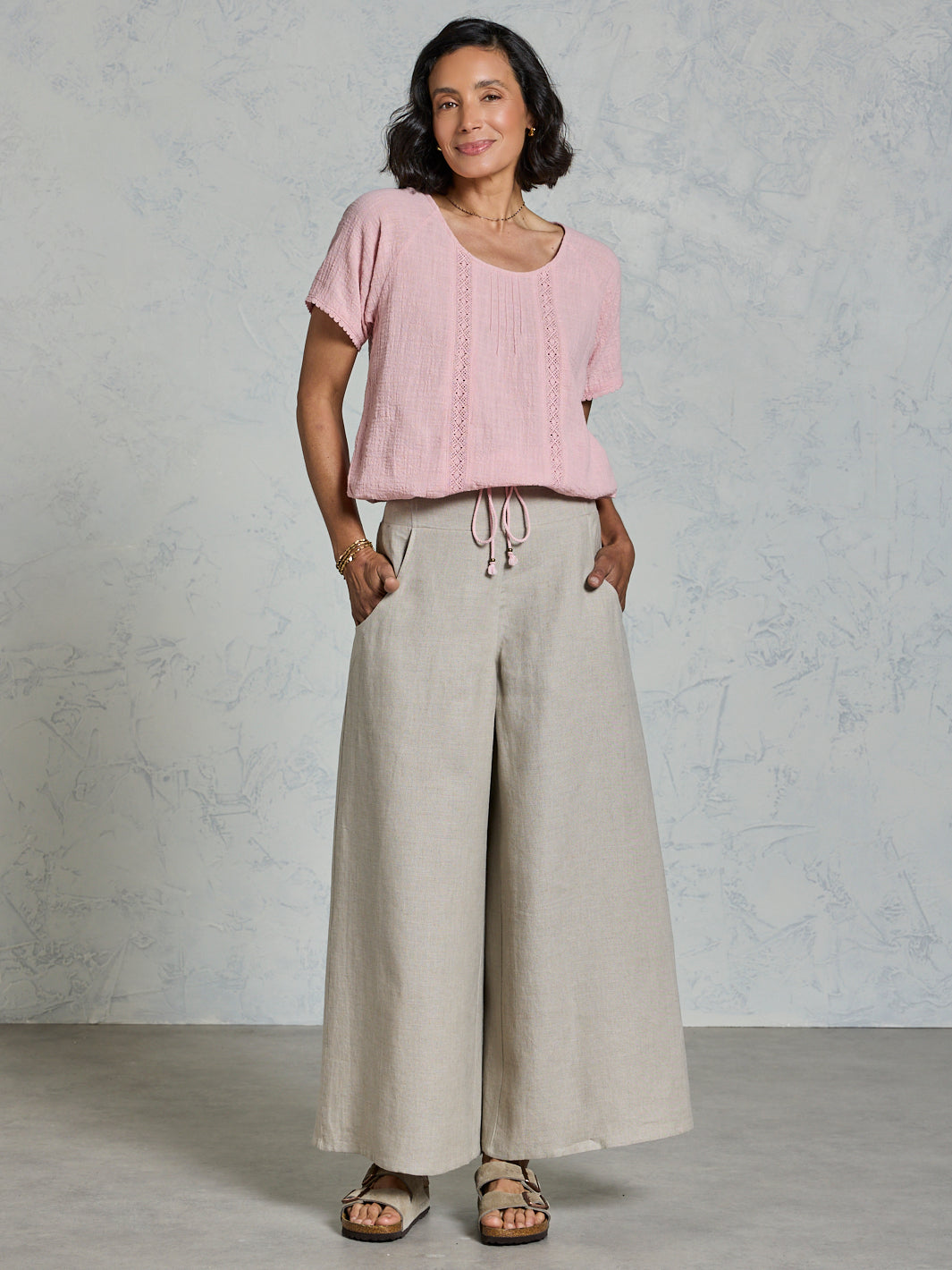 Woman wearing a pink top and beige wide-leg pants against a gray background