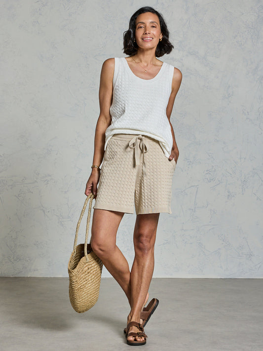 Woman wearing a white sleeveless top and beige shorts, holding a woven bag against a plain background