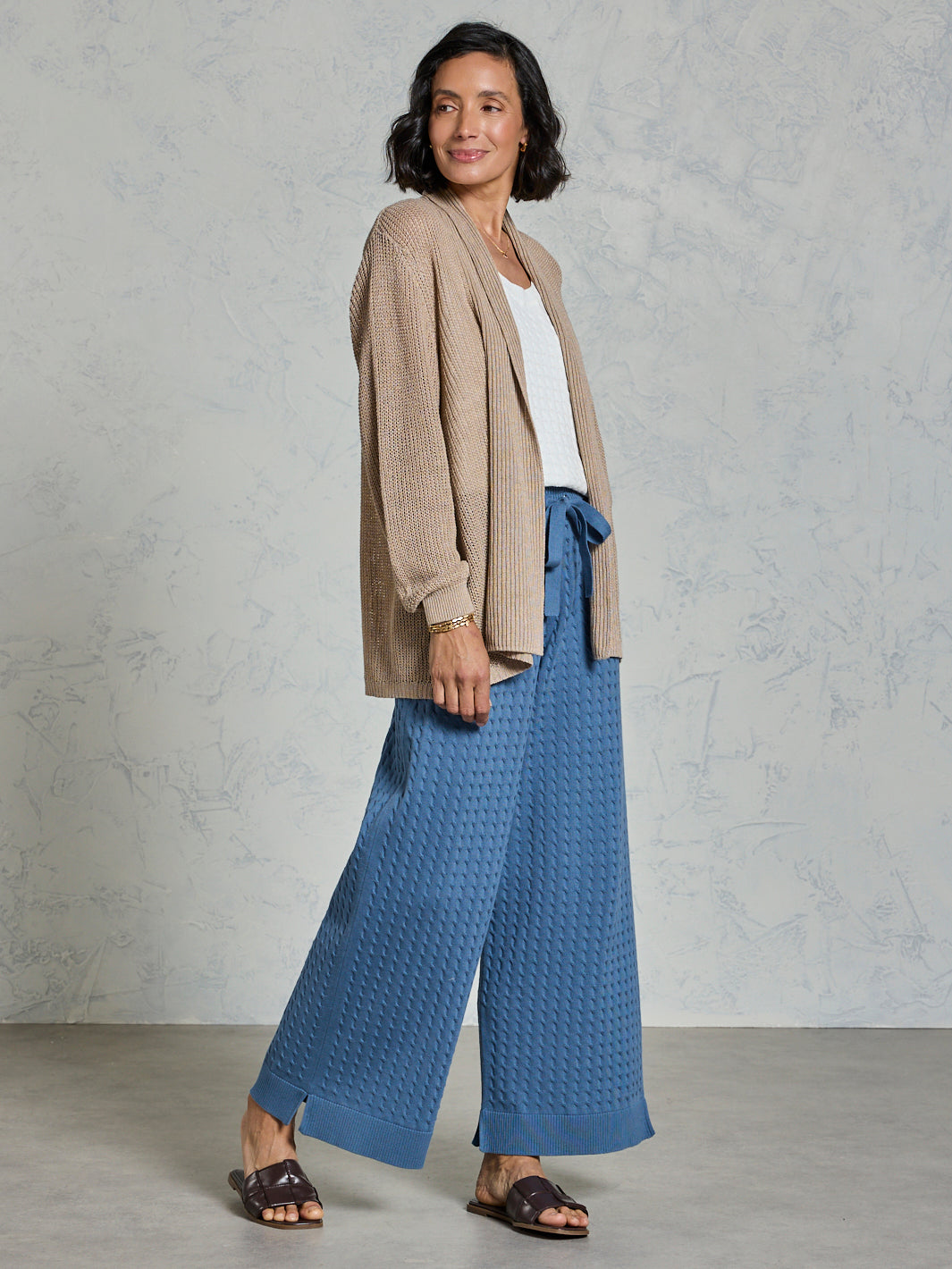 Woman wearing a beige cardigan and blue pants against a gray background