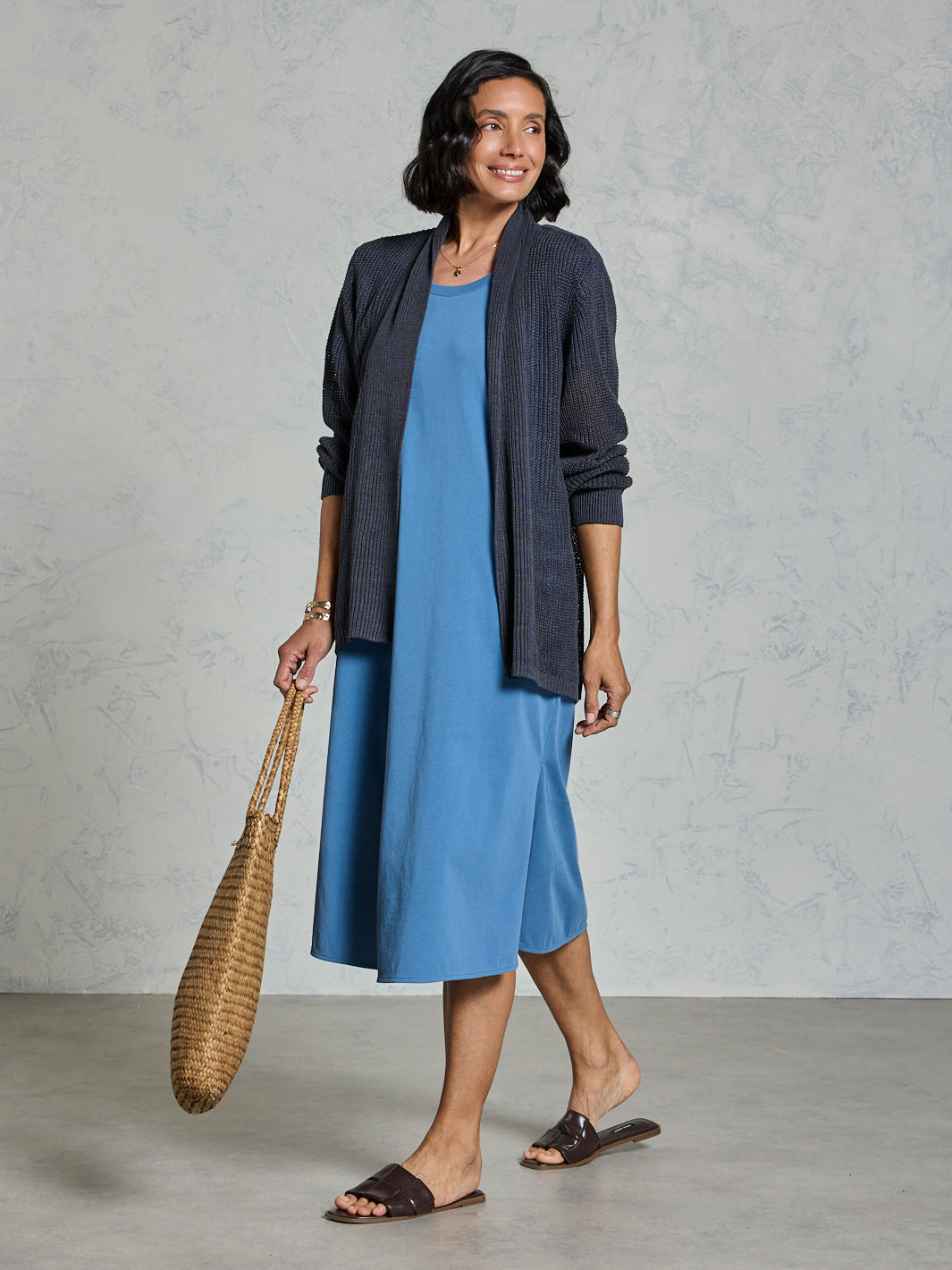 Woman wearing a blue dress and dark cardigan holding a woven object against a gray background
