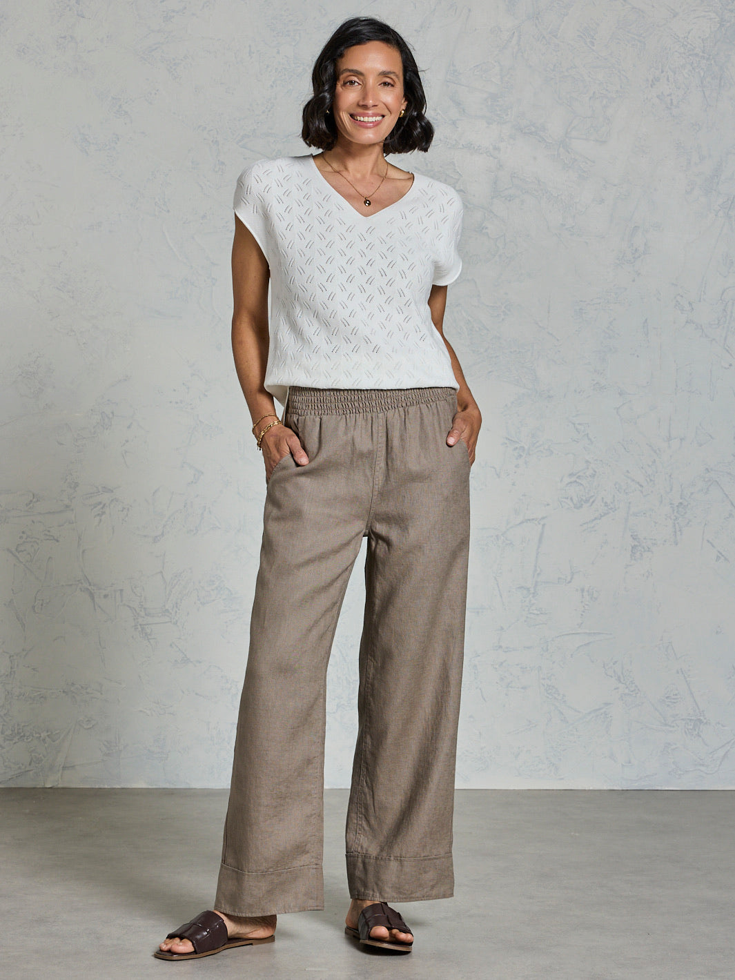 Woman wearing a white top and brown pants against a gray background