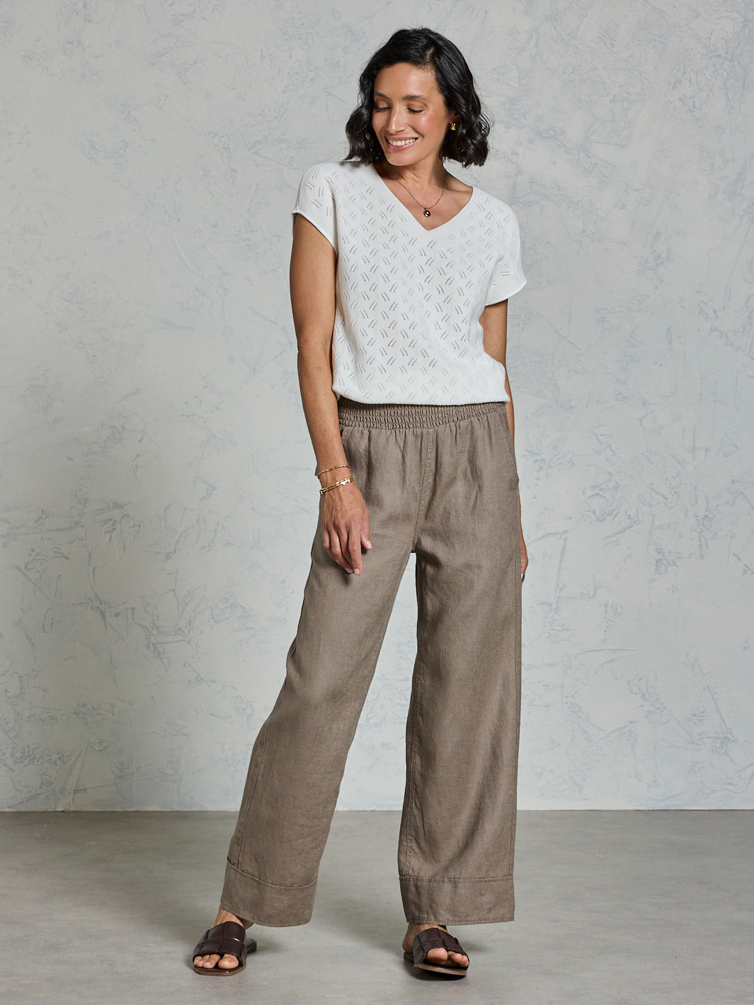 Woman wearing a white top and brown pants against a gray background