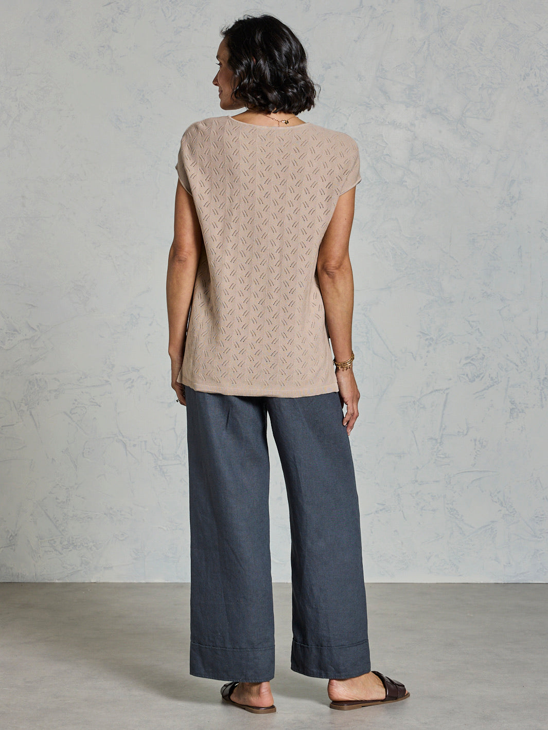 Woman wearing a beige sleeveless top and dark pants against a plain background