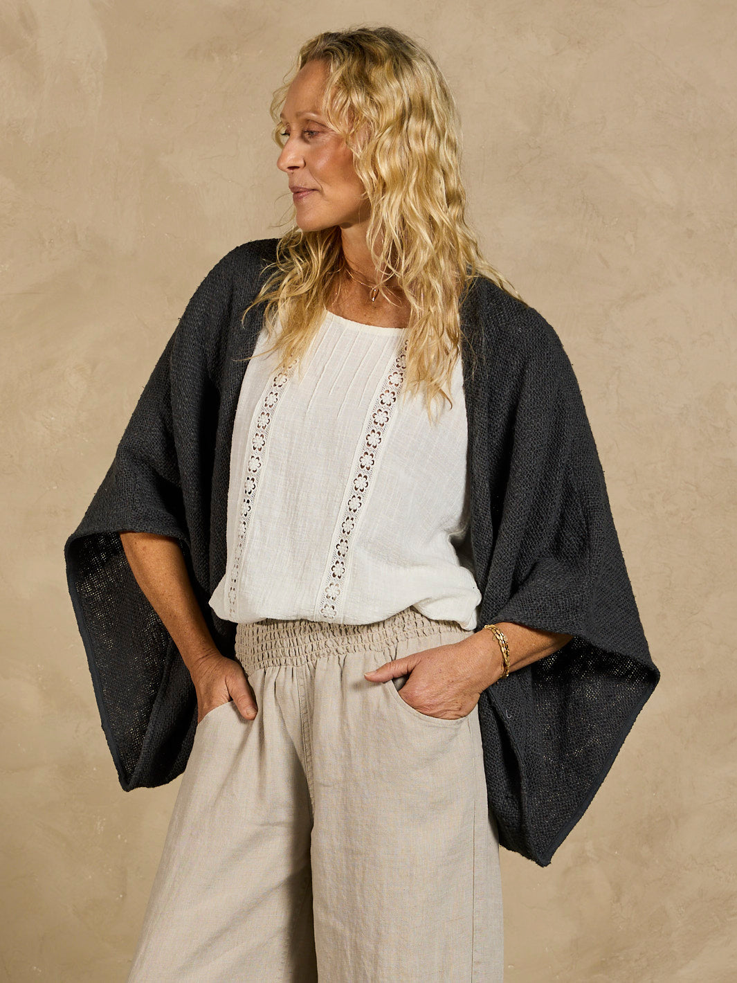 Woman wearing a black cardigan over a white top and beige pants against a beige background