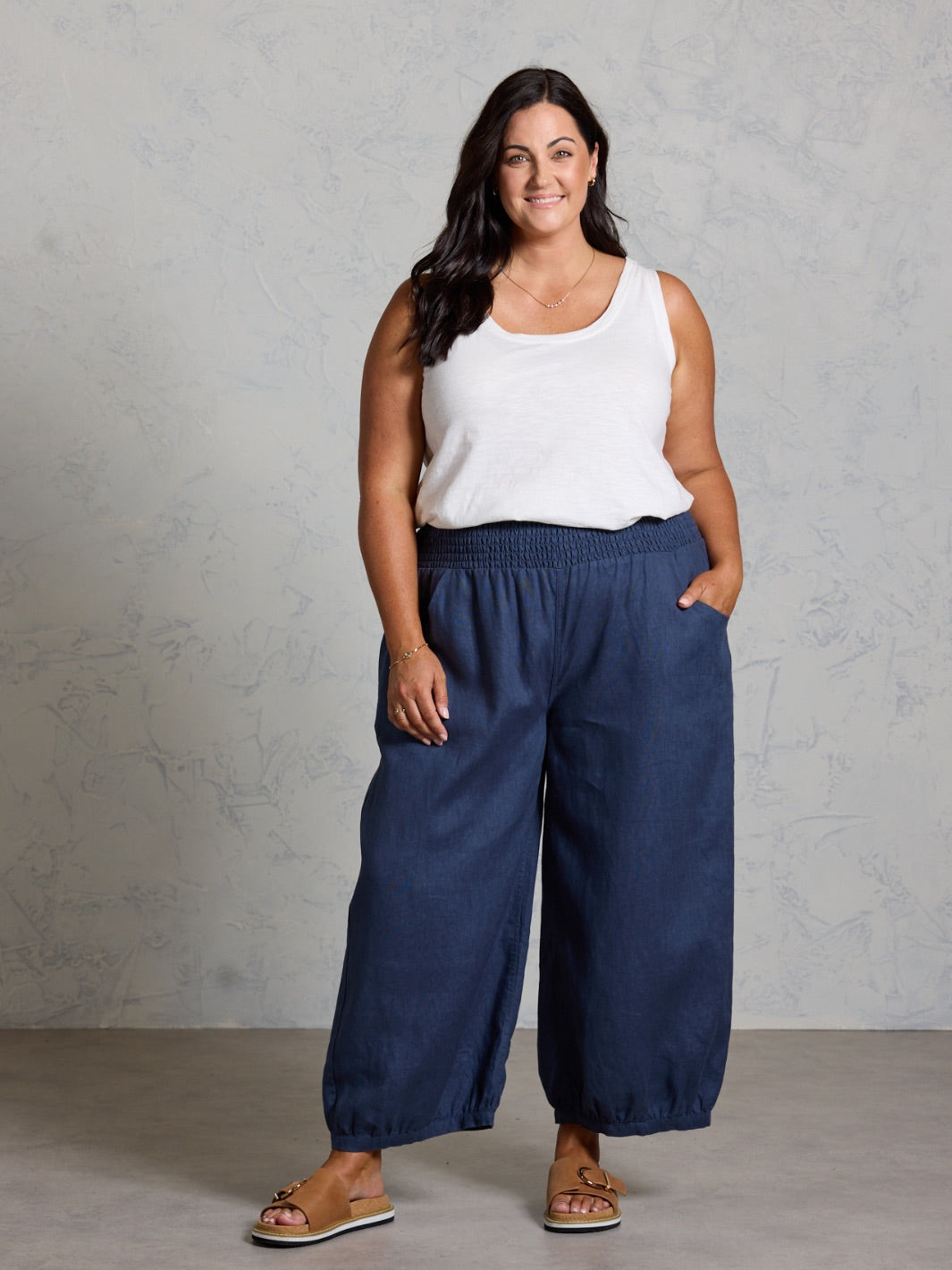 Woman wearing a white tank top and navy blue wide-leg pants against a gray background