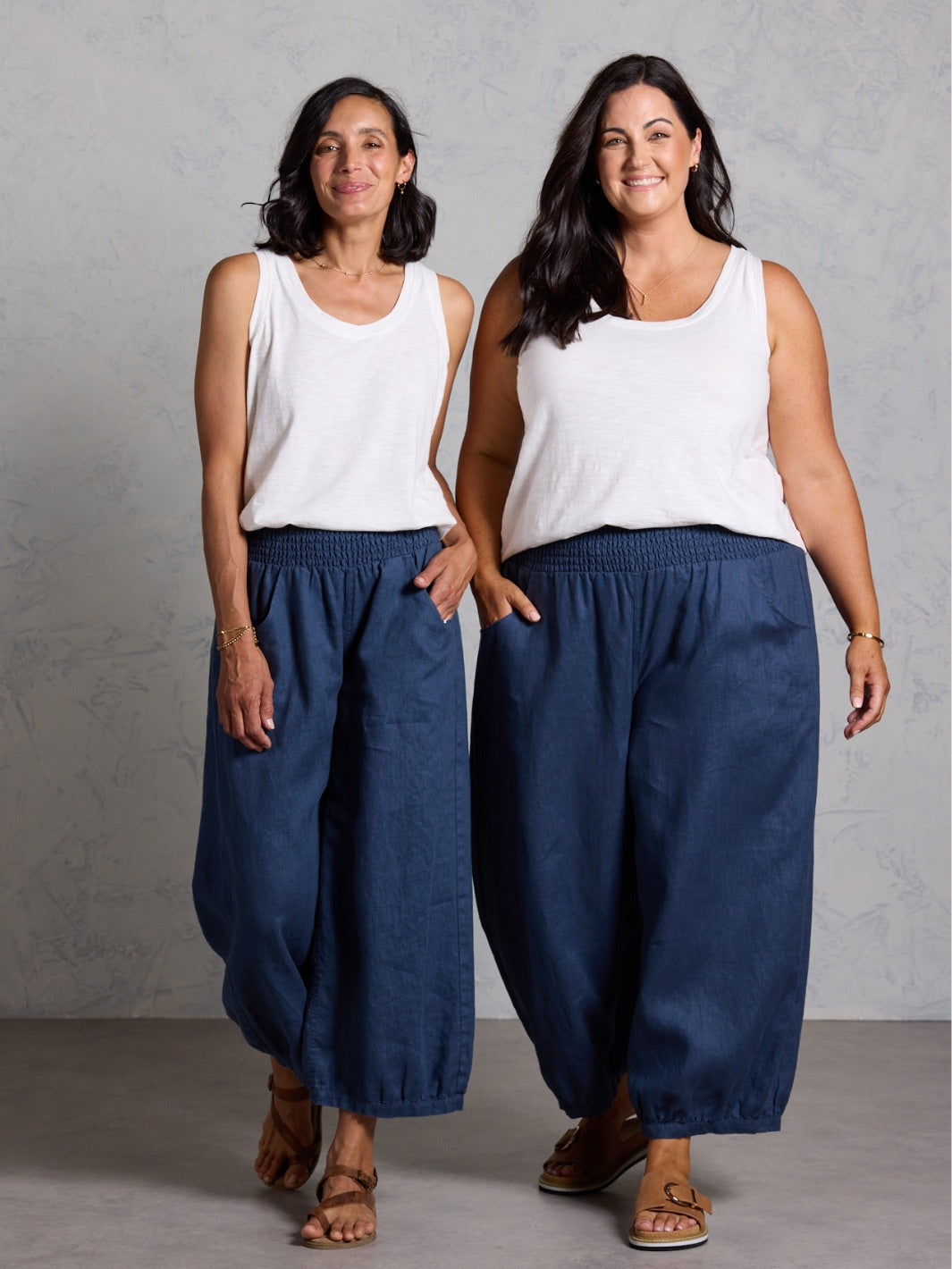 Two women wearing white tops and blue pants against a gray background