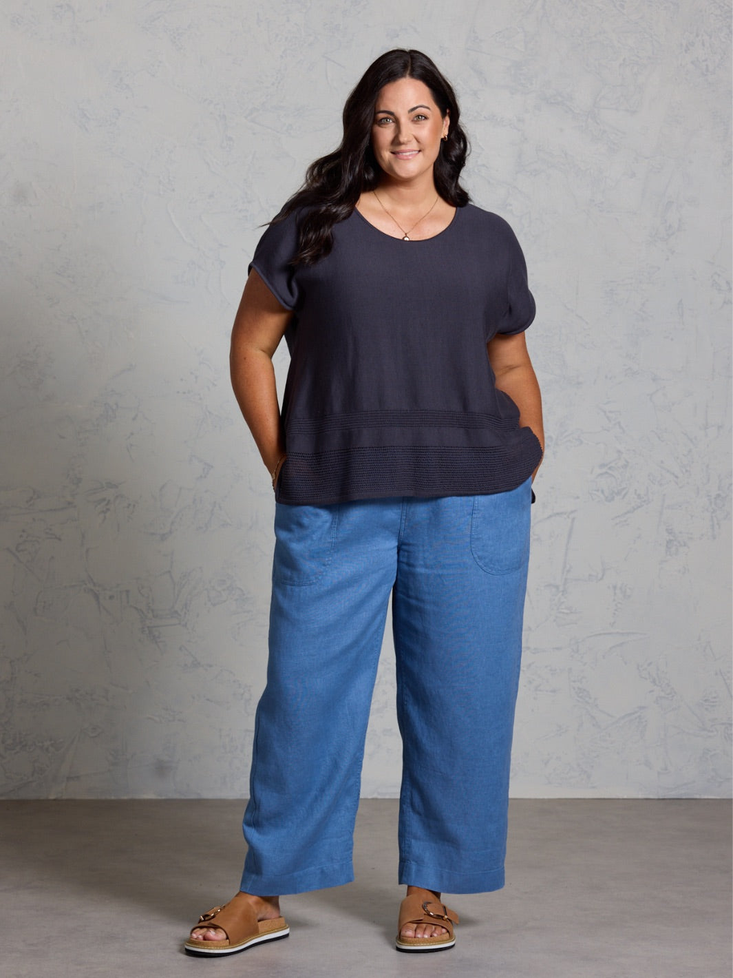 Woman wearing a black top and blue pants against a gray background