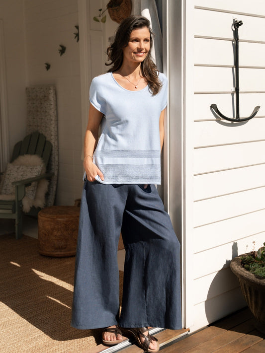 Woman standing on a porch wearing a light blue top and dark pants.
