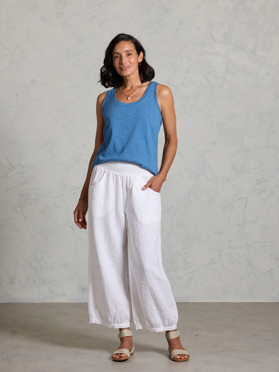 Woman wearing a blue tank top and white pants on a grey background.