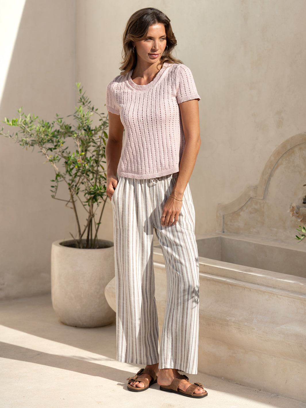 Model wearing the Agni Cotton Linen Knit Top in rosewater with striped linen drawstring pants in a sunlit stone courtyard.
