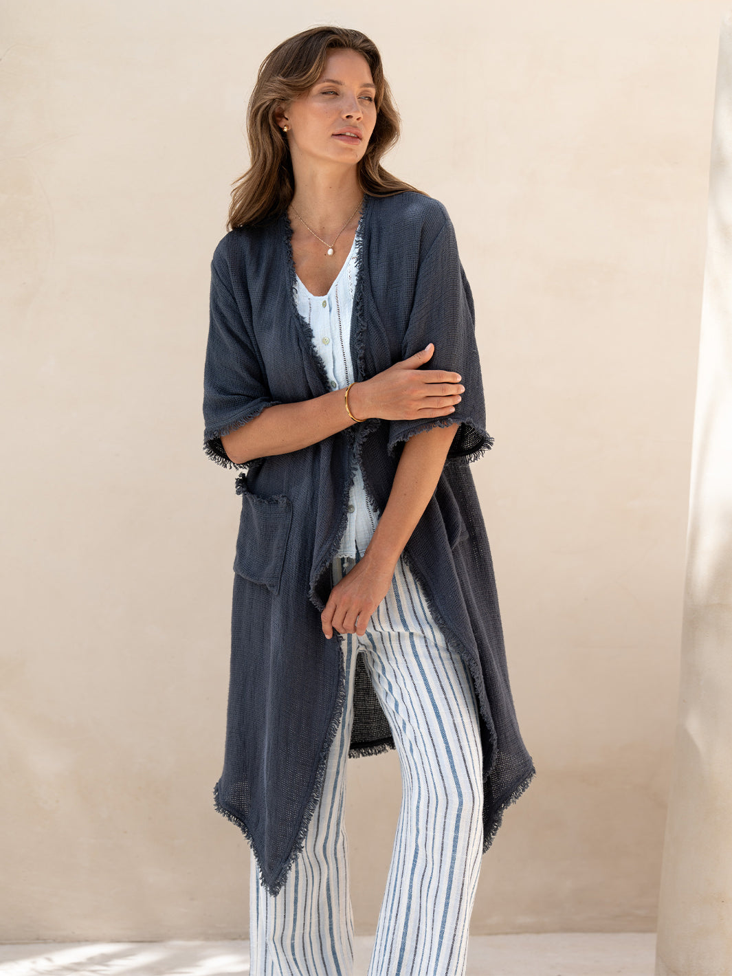 Model wearing the Laguna French Linen Kimono in charcoal, layered over a blue top and striped linen pants on sunlit stone steps.