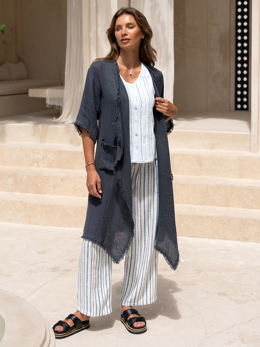 Model wearing the Laguna French Linen Kimono in charcoal, layered over a blue top and striped linen pants on sunlit stone steps.