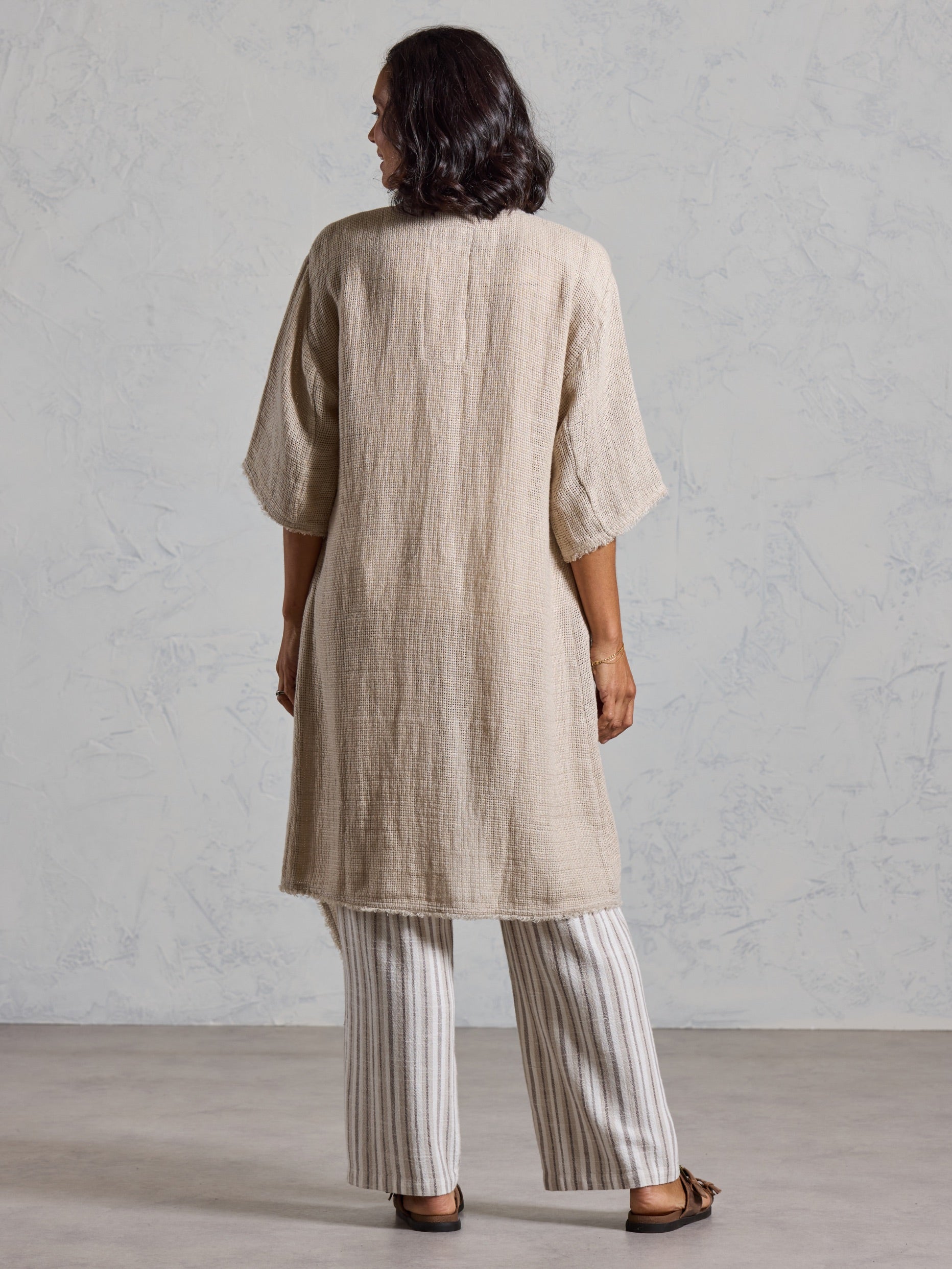 Back view of the Laguna French Linen Kimono in natural in motion, with striped linen pants in a sunlit interior.