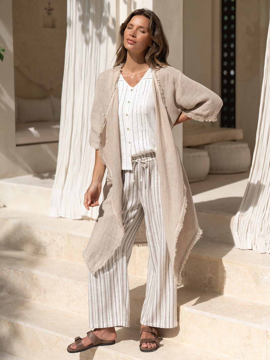 Model wearing the Laguna French Linen Kimono in natural with a white top and striped linen pants, on stone steps in dappled sunlight.